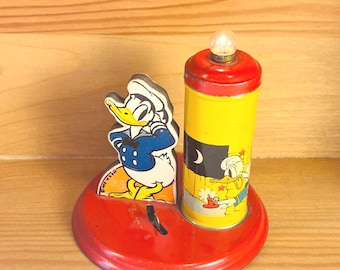May include: A vintage metal Donald Duck lamp featuring a red circular base, a cutout figure of Donald Duck in a blue sailor suit, and a yellow cylindrical light fixture topped with a small bulb, showing Donald Duck holding a candle.