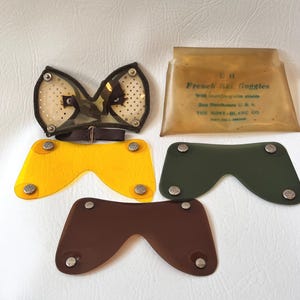 May include: Vintage French ski goggles with a clear lens and a brown strap, along with several colored replacement lenses in yellow, green, and brown. The goggles are in a clear plastic case with text.