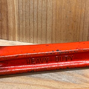 May include: Close-up of a red metal tool with the words "HEAVY DUTY" embossed on the side. The tool has a slightly worn appearance, suggesting it has been used. The background is a light brown wooden surface.