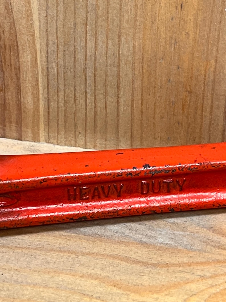 May include: Close-up of a red metal tool with the words "HEAVY DUTY" embossed on the side. The tool has a slightly worn appearance, suggesting it has been used. The background is a light brown wooden surface.