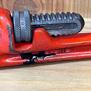 May include: Close-up of a red pipe wrench with a silver adjustable jaw. The wrench is marked with the number 8. The tool shows signs of wear and tear, with some black paint loss.