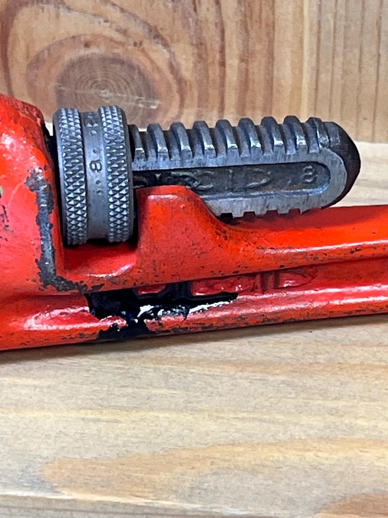 May include: Close-up of a red pipe wrench with a silver adjustable jaw. The wrench is marked with the number 8. The tool shows signs of wear and tear, with some black paint loss.