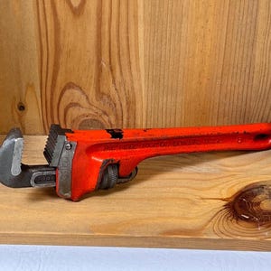 May include: A red pipe wrench with a black jaw, resting on a wooden surface. The wrench is a hand tool used for turning pipes or fittings. The tool is made of metal and has a textured handle.