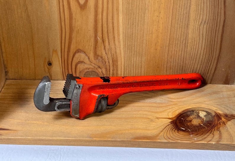 May include: A red pipe wrench with a black jaw, resting on a wooden surface. The wrench is a hand tool used for turning pipes or fittings. The tool is made of metal and has a textured handle.