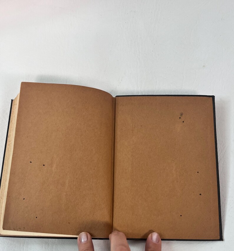 Puede incluir: Un libro abierto con p&aacute;ginas marrones y una cubierta negra. Las p&aacute;ginas est&aacute;n envejecidas, con peque&ntilde;as manchas oscuras. El libro est&aacute; sostenido abierto por dedos, con las p&aacute;ginas ligeramente curvadas. El fondo es una superficie blanca lisa.