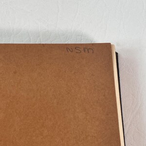 Puede incluir: Primer plano de un objeto rectangular marr&oacute;n con las letras manuscritas "NSM" en la esquina superior derecha. El objeto parece ser un trozo de madera o cart&oacute;n, posiblemente la parte posterior de un art&iacute;culo enmarcado. El fondo es una superficie blanca texturizada.