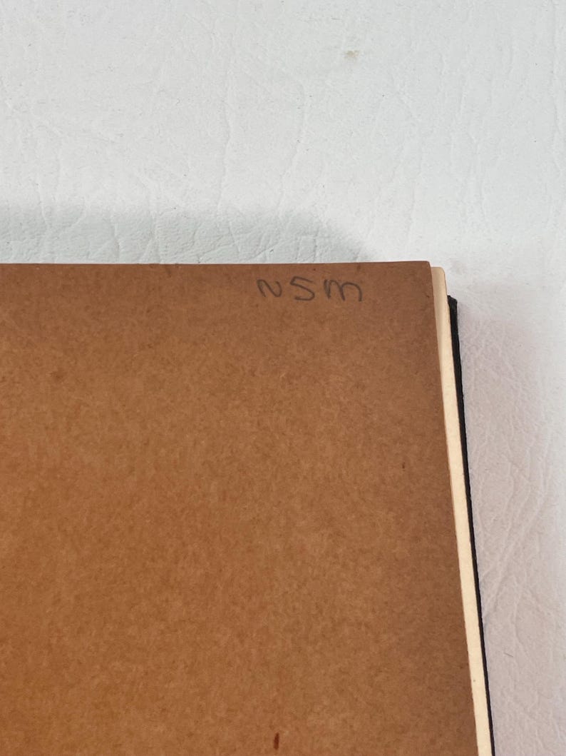 Puede incluir: Primer plano de un objeto rectangular marr&oacute;n con las letras manuscritas "NSM" en la esquina superior derecha. El objeto parece ser un trozo de madera o cart&oacute;n, posiblemente la parte posterior de un art&iacute;culo enmarcado. El fondo es una superficie blanca texturizada.