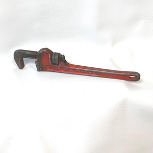 May include: A red pipe wrench with a long handle and adjustable jaws. The wrench is made of metal and has a textured grip. The text "PAT 1597807 14" is visible on the handle.