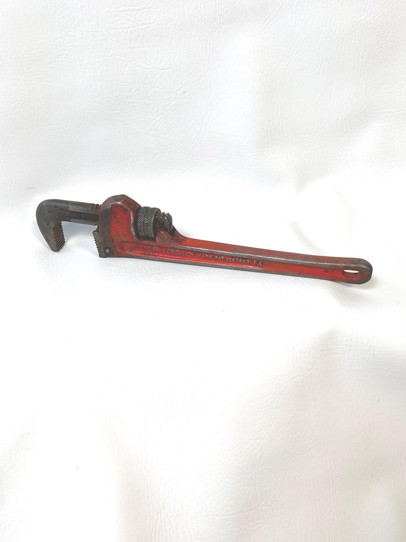 May include: A red pipe wrench with a long handle and adjustable jaws. The wrench is made of metal and has a textured grip. The text "PAT 1597807 14" is visible on the handle.