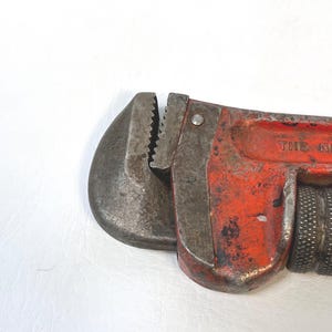 May include: Close-up of a vintage red and gray pipe wrench. The wrench has a textured handle and adjustable jaws. The metal shows signs of wear and tear, indicating its age and use. The wrench is a tool used for plumbing and other mechanical tasks.
