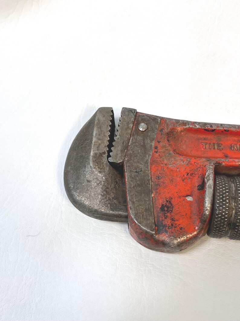 May include: Close-up of a vintage red and gray pipe wrench. The wrench has a textured handle and adjustable jaws. The metal shows signs of wear and tear, indicating its age and use. The wrench is a tool used for plumbing and other mechanical tasks.