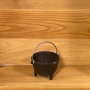 May include: A small, black, cast iron cauldron with a rounded body, three short legs, and a thin, arched metal handle attached to the sides. The interior is hollow and dark, and the exterior surface has a matte, textured finish.