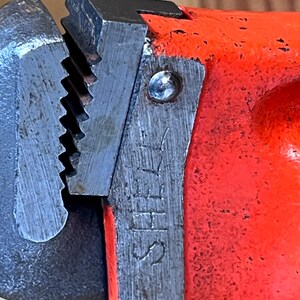 May include: Close-up of a red and silver pipe wrench. The wrench has a textured, toothed jaw and a bright red handle. The word "SHELL" is stamped on the metal. The tool appears well-used.