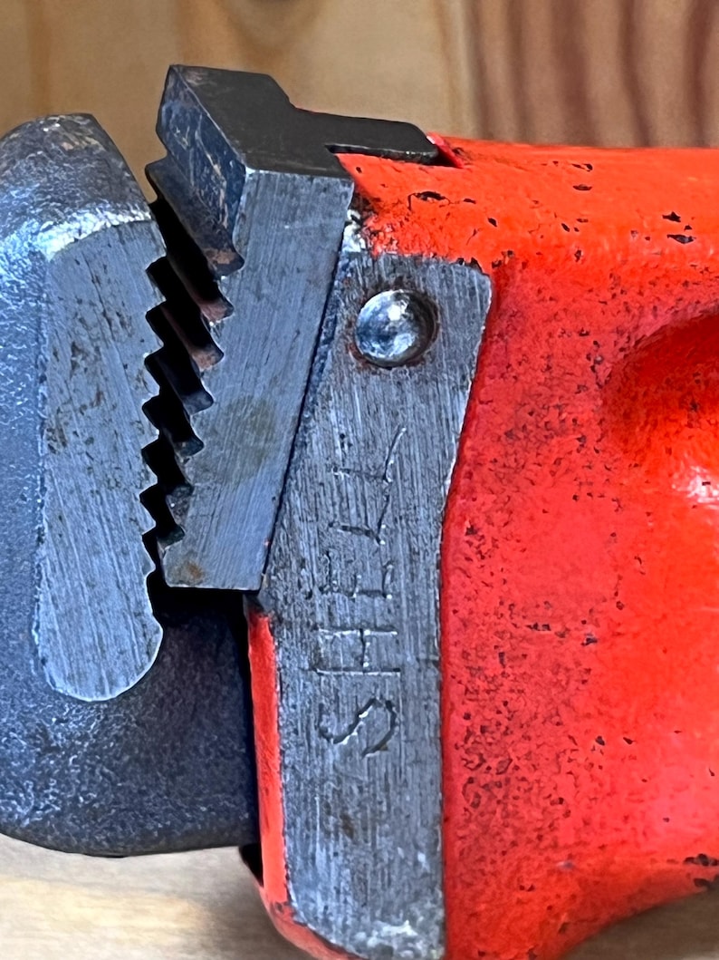 May include: Close-up of a red and silver pipe wrench. The wrench has a textured, toothed jaw and a bright red handle. The word "SHELL" is stamped on the metal. The tool appears well-used.