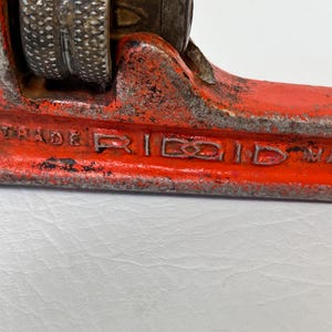 May include: Close-up of a vintage red and silver pipe wrench. The wrench has a textured handle and the word "TRADE RIDGID" is visible. The tool shows signs of wear and tear, indicating its age and use. A classic plumbing tool.