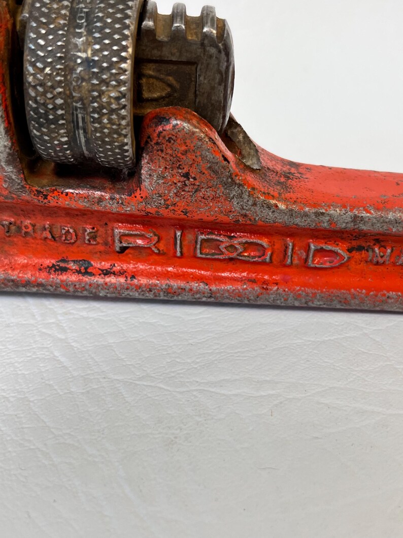 May include: Close-up of a vintage red and silver pipe wrench. The wrench has a textured handle and the word "TRADE RIDGID" is visible. The tool shows signs of wear and tear, indicating its age and use. A classic plumbing tool.