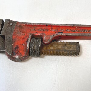 May include: Close-up of a vintage red and silver pipe wrench. The wrench has a textured handle and adjustable jaws. The text "THE RIDGE TOOL CO ELYRIA OHIO U.S.A." is visible on the wrench.