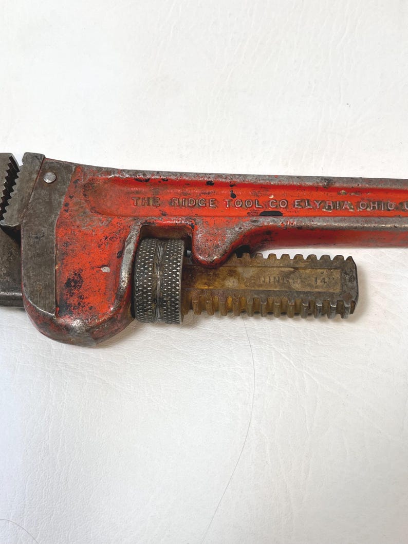 May include: Close-up of a vintage red and silver pipe wrench. The wrench has a textured handle and adjustable jaws. The text "THE RIDGE TOOL CO ELYRIA OHIO U.S.A." is visible on the wrench.