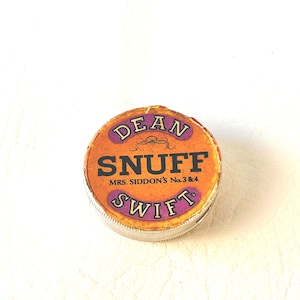 May include: A round metal tin with a worn orange label featuring the text DEAN SWIFT SNUFF and MRS. SIDDON'S No. 3 & 4 in black and purple lettering. The container has a metallic silver rim and shows signs of age and wear on the edges of the lid.