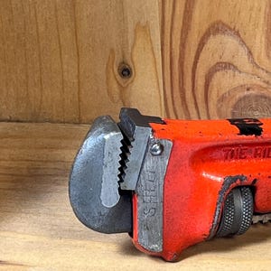 May include: Close-up of a red and black pipe wrench. The wrench has a steel jaw and a textured handle for grip. The word "STEEL" is visible on the side of the jaw. The tool is designed for gripping and turning pipes.