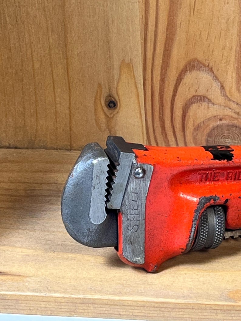 May include: Close-up of a red and black pipe wrench. The wrench has a steel jaw and a textured handle for grip. The word "STEEL" is visible on the side of the jaw. The tool is designed for gripping and turning pipes.