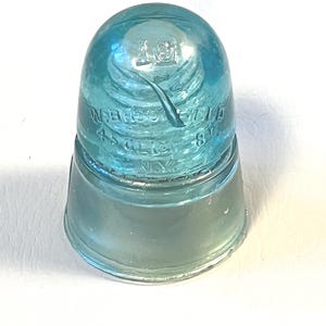 May include: A vintage, light blue glass insulator with a rounded top and a flared base. The insulator has embossed text that reads "WBCO FIELD 45 G118 NY". The glass has a slight swirl pattern and is translucent.