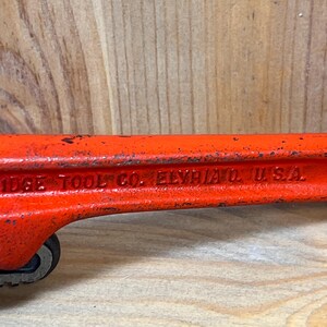 May include: Close-up of a vintage orange pipe wrench. The text "RIDGE TOOL CO. ELYRIA O. U.S.A." is visible on the side. The wrench shows signs of wear, with some paint chipping, indicating its age and use.