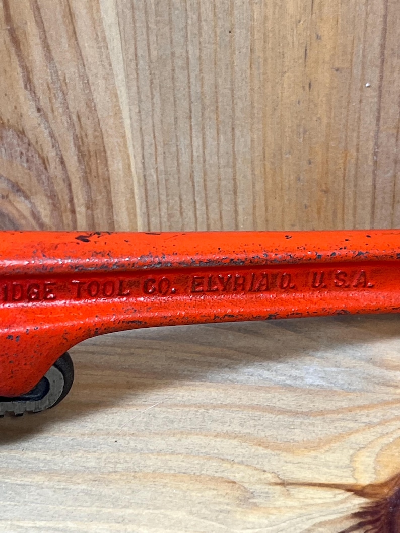May include: Close-up of a vintage orange pipe wrench. The text "RIDGE TOOL CO. ELYRIA O. U.S.A." is visible on the side. The wrench shows signs of wear, with some paint chipping, indicating its age and use.
