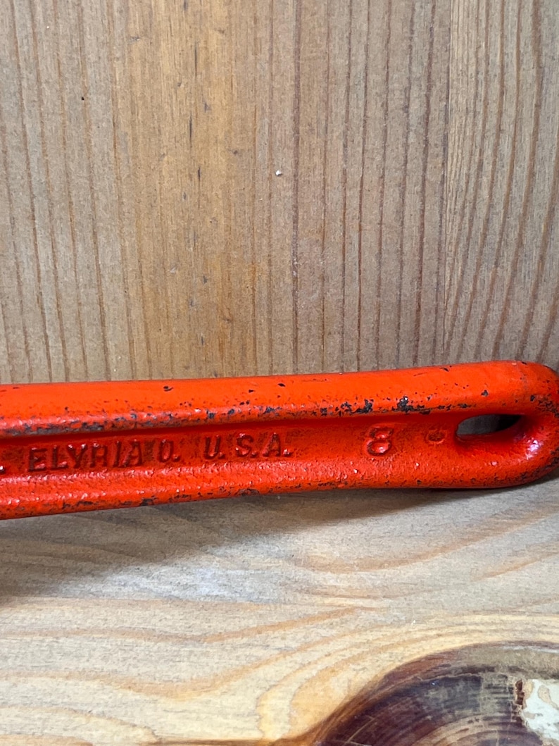 May include: Close-up of a vintage orange wrench with the text "ELYRIA O. USA" and the number "8" embossed on the side. The tool has a loop at the end and shows signs of wear.