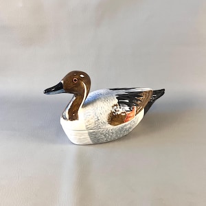 May include: A ceramic duck figurine featuring a brown head, a black beak, and a white body with grey and brown textured feather details. The duck has a horizontal seam across the middle of its body, indicating a lidded container design.