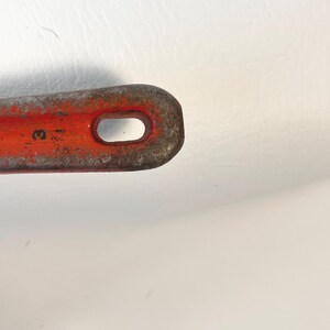 May include: Close-up of a rusty, orange metal tool with a rounded end and an oval-shaped hole. The tool has a worn appearance, with visible signs of age and use. The number "3" is partially visible on the tool.