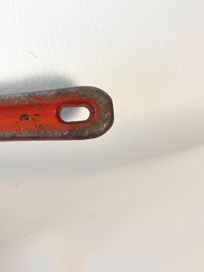 May include: Close-up of a rusty, orange metal tool with a rounded end and an oval-shaped hole. The tool has a worn appearance, with visible signs of age and use. The number "3" is partially visible on the tool.