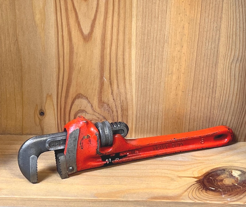 May include: A heavy-duty, adjustable pipe wrench with a bright red handle and a black jaw. The wrench is lying on a wooden surface, with the text "HEAVY DUTY" visible on the handle.