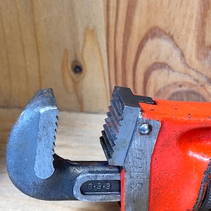May include: Close-up of a red and black pipe wrench. The wrench has a black jaw with teeth and a red handle. The word "STEEL" is visible on the side of the wrench.