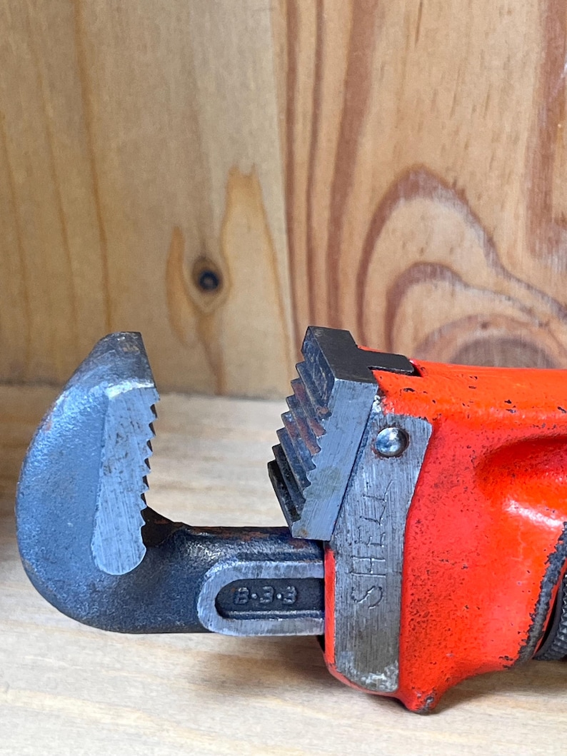 May include: Close-up of a red and black pipe wrench. The wrench has a black jaw with teeth and a red handle. The word "STEEL" is visible on the side of the wrench.
