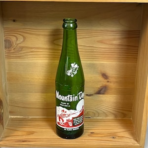 May include: A vintage green glass Mountain Dew bottle with white and red label graphics. The label features a cowboy illustration and the text "Mountain Dew" and "10 fluid ounces". The bottle is displayed inside a wooden box.