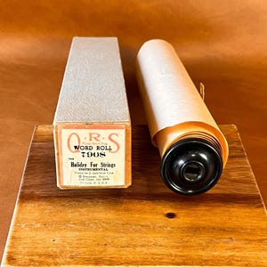 May include: A vintage Q.R.S. Word Roll 7908 piano roll titled Holiday For Strings, played by J. Lawrence Cook. The item includes a rectangular cardboard box with a light gray textured finish and a rolled paper sheet with a black plastic end cap.