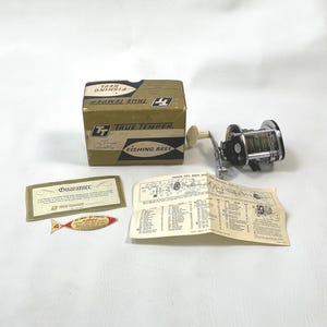 Puede incluir: Juego de carrete de pesca True Temper vintage. El juego incluye un carrete de pesca plateado y negro, una caja de cartón con las palabras "True Temper Fishing Reel", una tarjeta de garantía y una etiqueta con forma de pez.