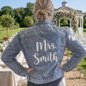May include: A light blue denim jacket with pearl embellishments on the shoulders and the text "Mrs. Smith" in white script. The jacket is part of a bridal ensemble, perfect for a wedding.