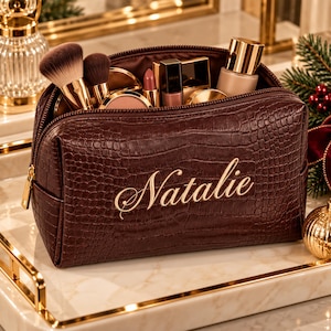 May include: A dark brown, crocodile-textured make-up bag featuring the name Natalie embroidered in gold script on the front. The open bag is filled with various cosmetic items, including make-up brushes, a lipstick, and a bottle of foundation.