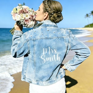 May include: Light blue denim jacket embellished with pearls, featuring the text "Mrs Smith" on the back. The jacket has long sleeves and is worn on a beach. The date "26.06.22" is printed on the collar.