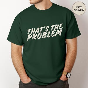 May include: A dark green t-shirt with the text "THAT'S THE PROBLEM" in white, stylized lettering. The shirt is short-sleeved and appears to be made of a soft material. A watch is visible on the wrist.