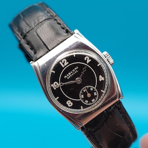 May include: A vintage Marconi Special wristwatch with a black face and a small seconds dial. The watch has a silver-coloured rectangular case and a black leather strap. The watch face displays the numbers 12, 2, 4, and 8.