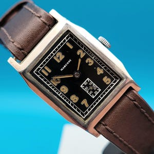 May include: A vintage Marconi wristwatch with a rectangular black face and gold numerals. The watch has a brown leather strap and a silver-coloured case. The watch face displays the Marconi brand name and a small seconds dial.
