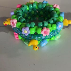 May include: A handmade kandi bracelet featuring green beads, adorned with pink, blue, and purple flower accents. Yellow duck charms with red beaks are also attached, creating a whimsical and colorful accessory.