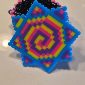 May include: A colorful beaded bracelet with a blue, star-shaped charm. The charm features a pixelated design in shades of yellow, pink, and purple. The bracelet itself is made of black, pink, and purple beads, creating a vibrant accessory.
