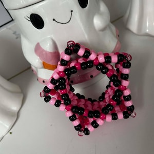 Könnte beinhalten: Ein sternförmiges Armband aus Perlen in Rosa- und Schwarztönen. Das Armband besteht aus kleinen, runden Perlen, die zu einer geometrischen Form aufgereiht sind. Das Armband liegt auf einer weißen Oberfläche.