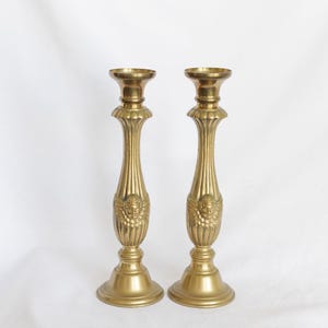 May include: Two gold-colored, vintage candle holders. Each candlestick has a round base, a fluted stem, and a detailed cherub design. The top of each holder is designed to hold a candle. The candle holders are approximately 10 inches tall.