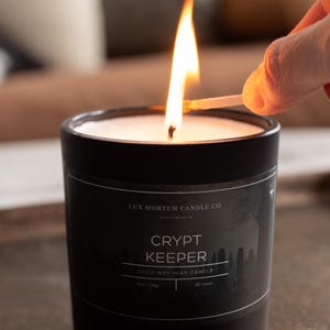 May include: A black candle with a white wax interior is lit with a match. The candle has a label that reads "CRYPT KEEPER" and "COCO-SOY WAX CANDLE". The candle is 8 oz / 225g and has a burn time of 35 hours. The candle is in a black ceramic container.