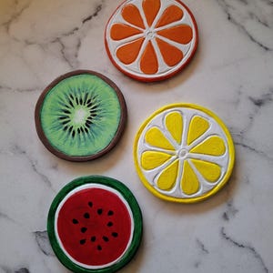 Op de afbeelding: Vier kleurrijke, ronde onderzetters met fruitontwerpen. De onderzetters tonen een sinaasappelschijf, kiwi, citroen en watermeloen. De oranje onderzetter heeft oranje en witte segmenten met een rode rand. De kiwi heeft groene en bruine tinten. De citroen heeft gele en witte segmenten.
